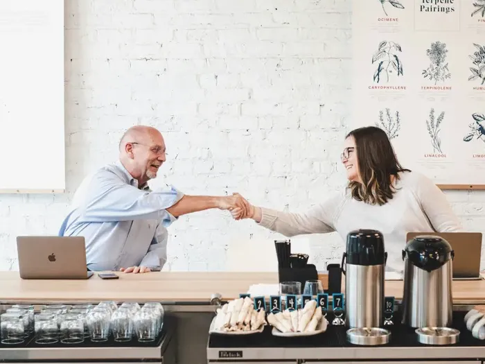 Two colleagues smiling and shaking hands across a cafe counter during casual business meeting with laptops on table